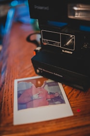 A Polaroid camera is placed on a wooden surface, with a printed photo lying beside it. The photo features a person in a red garment making a hand gesture. The surrounding scene is softly focused, giving a cozy and nostalgic atmosphere.