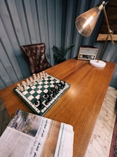 A wooden desk features a chess board with pieces set up for a game. An old-fashioned radio sits on the desk next to a large, angled lamp with a warm bulb. A brown, tufted leather chair is placed behind the desk. A newspaper with text and images is partially visible in the foreground. The room has blue panel walls and a plant placed near the chair.
