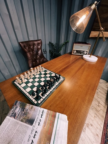A wooden desk features a chess board with pieces set up for a game. An old-fashioned radio sits on the desk next to a large, angled lamp with a warm bulb. A brown, tufted leather chair is placed behind the desk. A newspaper with text and images is partially visible in the foreground. The room has blue panel walls and a plant placed near the chair.