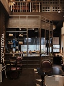 The interior of a cozy cafe with wooden and metal elements, featuring a well-organized bar with shelves holding various bottles and decor. The seating area includes dark, comfortable chairs and tables, while the overall setting has a warm, inviting ambiance.