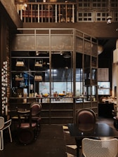 The interior of a cozy cafe with wooden and metal elements, featuring a well-organized bar with shelves holding various bottles and decor. The seating area includes dark, comfortable chairs and tables, while the overall setting has a warm, inviting ambiance.