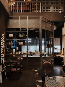 The interior of a cozy cafe with wooden and metal elements, featuring a well-organized bar with shelves holding various bottles and decor. The seating area includes dark, comfortable chairs and tables, while the overall setting has a warm, inviting ambiance.