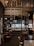 The interior of a cozy cafe with wooden and metal elements, featuring a well-organized bar with shelves holding various bottles and decor. The seating area includes dark, comfortable chairs and tables, while the overall setting has a warm, inviting ambiance.