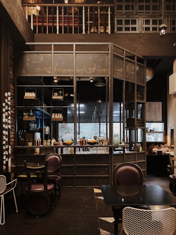The interior of a cozy cafe with wooden and metal elements, featuring a well-organized bar with shelves holding various bottles and decor. The seating area includes dark, comfortable chairs and tables, while the overall setting has a warm, inviting ambiance.