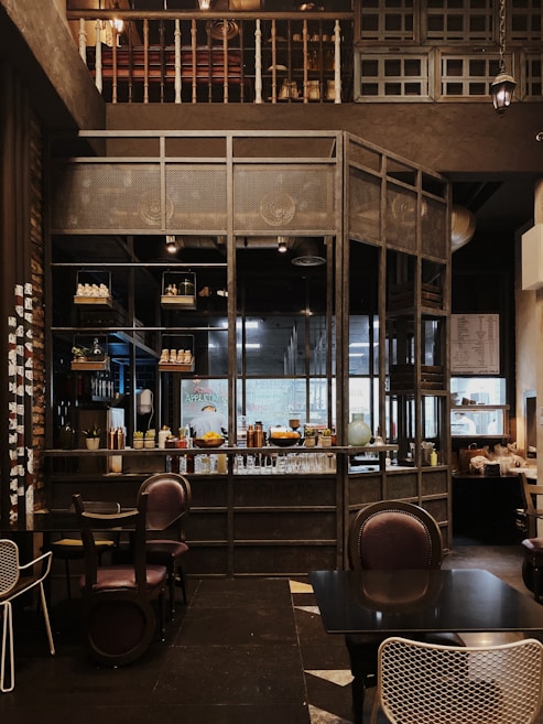 The interior of a cozy cafe with wooden and metal elements, featuring a well-organized bar with shelves holding various bottles and decor. The seating area includes dark, comfortable chairs and tables, while the overall setting has a warm, inviting ambiance.