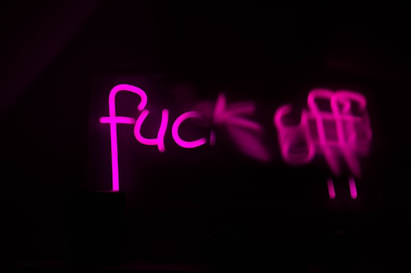 Bold pink neon letters illuminate against a dark background, displaying an explicit phrase partially obscured by blur.