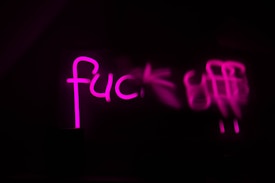 Bold pink neon letters illuminate against a dark background, displaying an explicit phrase partially obscured by blur.