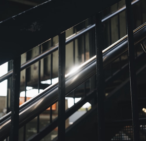 The image features a set of stairs with metallic railings, with sunlight glinting off the polished surface. The structure appears to be an outdoor staircase, with shadows and a slightly industrial feel due to visible metalwork and surrounding architecture.