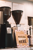 Two commercial coffee grinders filled with coffee beans stand on a counter next to several syrup bottles and a wooden sign with floral decorations that reads 'Alfred Matcha Served Here'.