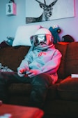A person wearing a space helmet and a grey hoodie is sitting on a brown sofa, playing a video game with a controller in hand. The room has a relaxed and slightly cluttered atmosphere with stuffed animals, a hat, and a drawing of a deer on the wall.