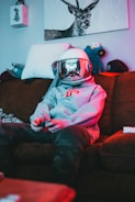 A person wearing a space helmet and a grey hoodie is sitting on a brown sofa, playing a video game with a controller in hand. The room has a relaxed and slightly cluttered atmosphere with stuffed animals, a hat, and a drawing of a deer on the wall.
