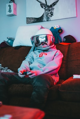 A person wearing a space helmet and a grey hoodie is sitting on a brown sofa, playing a video game with a controller in hand. The room has a relaxed and slightly cluttered atmosphere with stuffed animals, a hat, and a drawing of a deer on the wall.