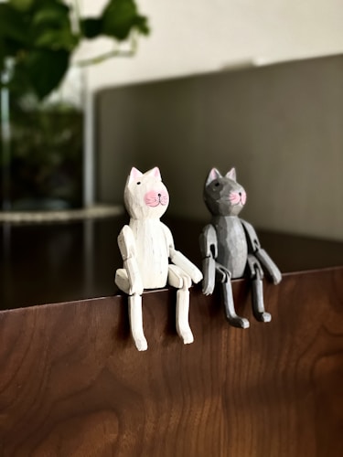 Two wooden cat figurines, one white and one gray, are sitting side by side on the edge of a wooden surface. The figurines have simple designs with joints to mimic sitting postures. In the background, there is a blurred view of a green plant, adding a natural touch to the scene.