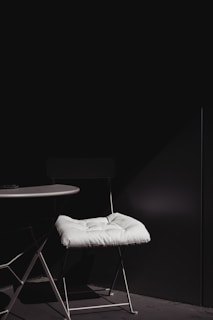 A minimalist setup featuring a white-cushioned chair positioned next to a round white table. The scene is dimly lit with a strong contrast between the white elements and the dark background, highlighting the simplicity and elegance of the furniture.