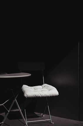 A minimalist setup featuring a white-cushioned chair positioned next to a round white table. The scene is dimly lit with a strong contrast between the white elements and the dark background, highlighting the simplicity and elegance of the furniture.