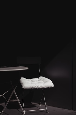 A minimalist setup featuring a white-cushioned chair positioned next to a round white table. The scene is dimly lit with a strong contrast between the white elements and the dark background, highlighting the simplicity and elegance of the furniture.