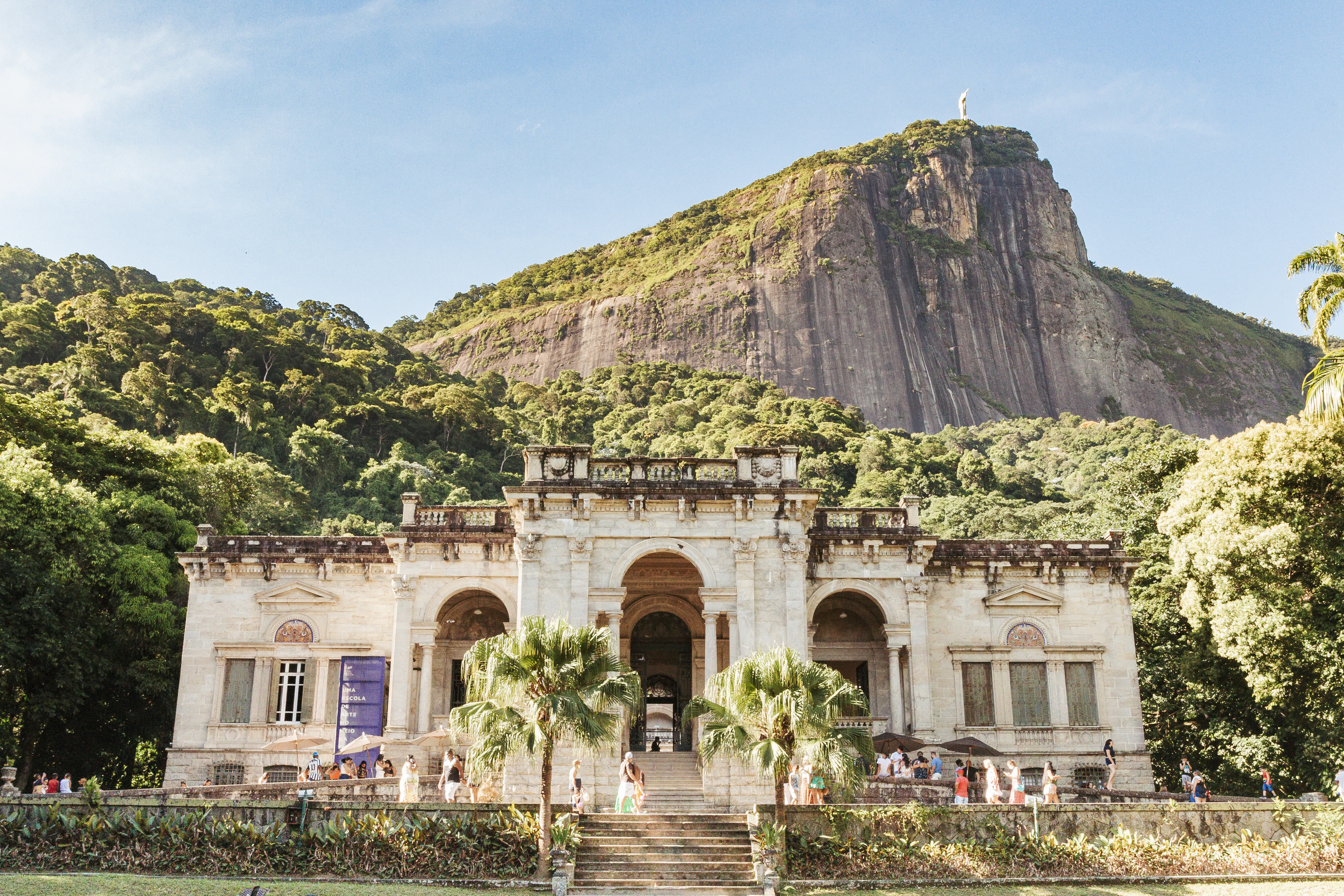 The colonnaded mansion at Parque Lage in Rio de Janeiro, palms in front of its arched entrance and the sheer granite wall of Corcovado rising out of the rainforest directly behind, Christ the Redeemer just visible on the summit