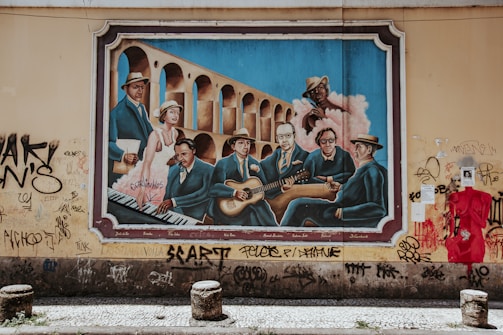 A vibrant mural painted on a wall depicting a group of musicians in blue suits, sitting in front of an arched structure. The artwork includes a pianist, guitarists, and various other figures, with an urban decay setting as graffiti surrounds the painting.