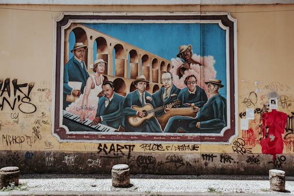 A vibrant mural painted on a wall depicting a group of musicians in blue suits, sitting in front of an arched structure. The artwork includes a pianist, guitarists, and various other figures, with an urban decay setting as graffiti surrounds the painting.
