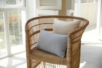 A wicker chair positioned in a bright, sunlit room with large windows. The chair is adorned with two cushions, one grey and one beige, adding a touch of comfort and style. The natural light creates soft shadows and an inviting atmosphere.