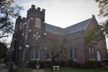 A historic brick church features Gothic architectural elements with pointed arches and large windows, surrounded by flowering shrubs and tall trees. Two prominent towers with louvered openings rise at the front, while the roof is steeply pitched.