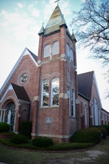 A traditional red brick church building with a prominent tower topped by a spire and cross. The structure features large arched windows and detailed masonry. Trees with sparse leaves are visible in the background, giving a sense of early spring or late fall.