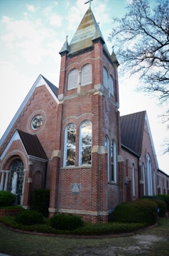 A traditional red brick church building with a prominent tower topped by a spire and cross. The structure features large arched windows and detailed masonry. Trees with sparse leaves are visible in the background, giving a sense of early spring or late fall.