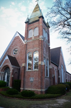 A traditional red brick church building with a prominent tower topped by a spire and cross. The structure features large arched windows and detailed masonry. Trees with sparse leaves are visible in the background, giving a sense of early spring or late fall.