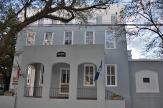 A large, light gray building with tall windows and a small entry porch supported by columns. The building has a sign indicating it is the Old Charleston Jail. An American flag and another flag are visible on poles near the entrance. Leafless trees frame the scene, and the sky is partly cloudy.