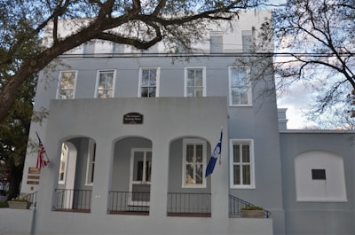 A large, light gray building with tall windows and a small entry porch supported by columns. The building has a sign indicating it is the Old Charleston Jail. An American flag and another flag are visible on poles near the entrance. Leafless trees frame the scene, and the sky is partly cloudy.