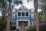 A two-story house with a combination of brick and blue siding features multiple windows and a small front porch. The roof has dormer windows with dark green trim, and there are two prominent palm trees in the foreground. Surrounding the house is a well-maintained garden with bushes and flowering plants.