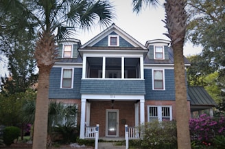 A two-story house with a combination of brick and blue siding features multiple windows and a small front porch. The roof has dormer windows with dark green trim, and there are two prominent palm trees in the foreground. Surrounding the house is a well-maintained garden with bushes and flowering plants.