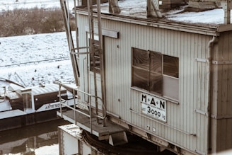 A waterfront industrial structure with a large sign indicating a weight limit of 3000 kg. The building appears weathered and aged, with metal railing and ladders visible. In the background, there is a barge partially covered with snow along the edge of a river.