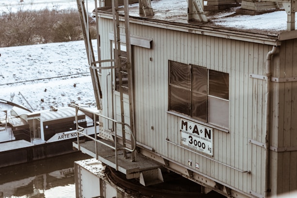 A waterfront industrial structure with a large sign indicating a weight limit of 3000 kg. The building appears weathered and aged, with metal railing and ladders visible. In the background, there is a barge partially covered with snow along the edge of a river.