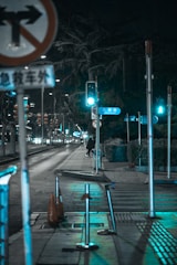 A nighttime urban street scene with traffic signs, traffic lights, and road markings. The street is illuminated by turquoise and white lights, with empty sidewalks and a person walking in the distance. The trees and decorative lanterns contribute to the urban setting.