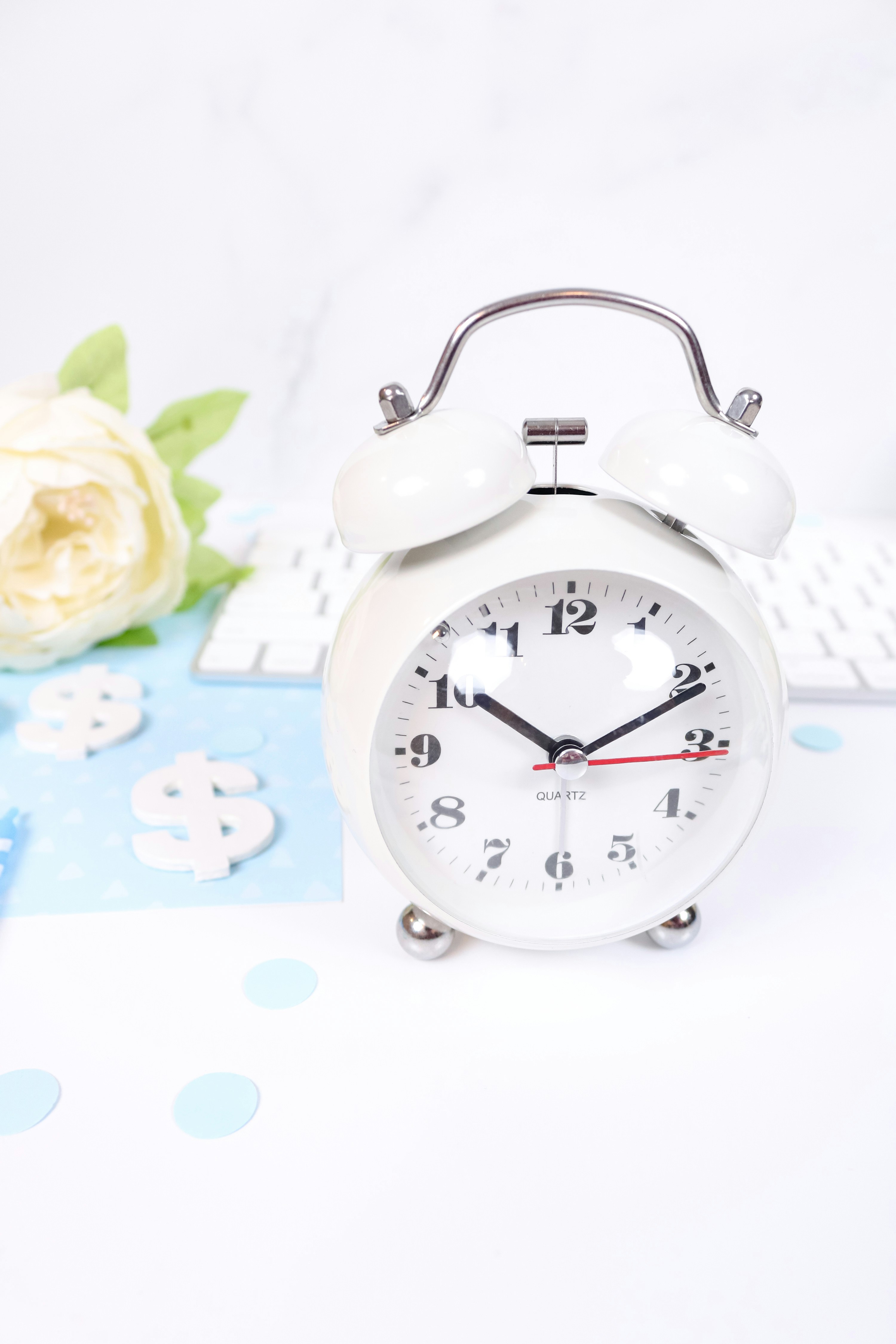 This feminine flat lay styled stock photo features a white desk with light blue and dark blue props like a keyboard, white flowers, markers, confetti, a clock for keeping track of time or representing productivity for entrepreneurs, and more.