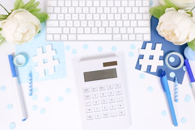 This feminine flat lay styled stock photo features a white desk with light blue and dark blue props like a keyboard, white flowers, markers, confetti, hashtags for social media, a calculator perfect for accountants or taxes for business, and more.