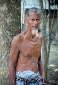 An older man stands in a natural outdoor setting, leaning against a tree. He is shirtless, wearing a patterned cloth around his waist. The background shows additional trees and a soft-focus environment, contributing to a rural atmosphere.