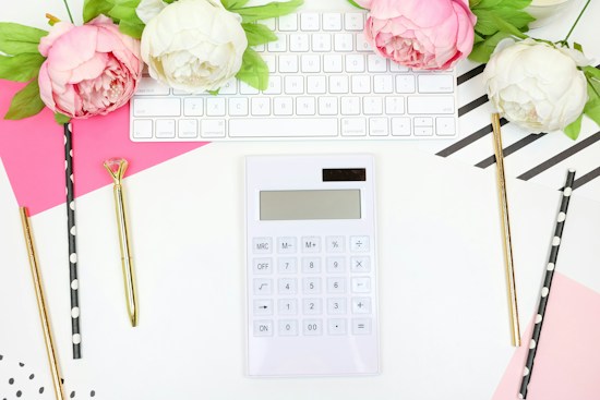 A bright, organized desk setup featuring a white calculator centered on a white surface. Surrounding the calculator are three gold pens, each with a distinct style, and black-and-white polka dot straws. Above, a white computer keyboard is paired with pink and white peony flowers, adding a soft, floral touch. The background includes pink and striped geometric patterns.