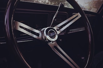 A classic Ford steering wheel with a three-spoke design, featuring a metallic finish and a central horn button displaying the Ford logo. The dashboard is visible in the background with some controls and a gear shift lever.