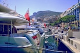 Luxurious yachts are docked at a marina with tall modern buildings and green hills in the background. The scene is bustling with people walking along the waterfront, and the area is lined with well-manicured trees.