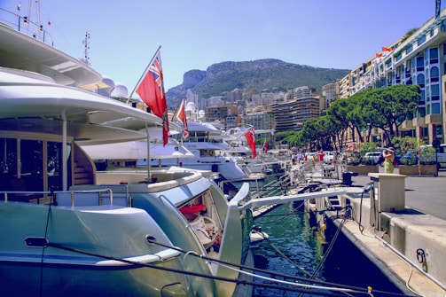 Luxurious yachts are docked at a marina with tall modern buildings and green hills in the background. The scene is bustling with people walking along the waterfront, and the area is lined with well-manicured trees.