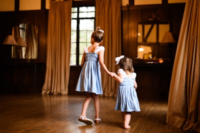 Two young girls wearing matching blue dresses are holding hands in a warmly lit room with wooden floors and beige curtains. A large window and a mirror above a table are visible in the background.