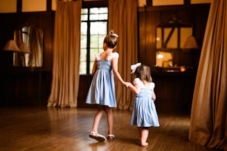 Two young girls wearing matching blue dresses are holding hands in a warmly lit room with wooden floors and beige curtains. A large window and a mirror above a table are visible in the background.