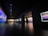 A dimly lit gallery features a series of large, vibrant landscape paintings on its walls. A person is walking through the space, examining the artwork, with more visitors visible further down the hallway. The gallery has a modern design with dark walls and a polished floor, enhanced by spotlighting focusing on the artworks.