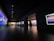 A dimly lit gallery features a series of large, vibrant landscape paintings on its walls. A person is walking through the space, examining the artwork, with more visitors visible further down the hallway. The gallery has a modern design with dark walls and a polished floor, enhanced by spotlighting focusing on the artworks.
