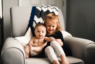 A young child with auburn hair is sitting on a soft, light-colored armchair, holding a baby. The child is wearing a black outfit with white pants, and the baby is dressed minimally. Behind them is a knit blanket with a zigzag pattern in blue, white, and gray.