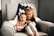 A young child with auburn hair is sitting on a soft, light-colored armchair, holding a baby. The child is wearing a black outfit with white pants, and the baby is dressed minimally. Behind them is a knit blanket with a zigzag pattern in blue, white, and gray.