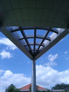 A modern architectural structure with a triangular canopy supported by a central pillar is seen against a clear blue sky with scattered clouds. The canopy has a grid-like pattern and the surroundings include greenery and the red-tiled roof of a building partially visible.
