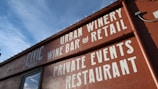 A building facade with bold white lettering on a reddish-brown wall advertising an urban winery, wine bar, restaurant, and space for private events. The text is prominently displayed against the backdrop of a clear blue sky.